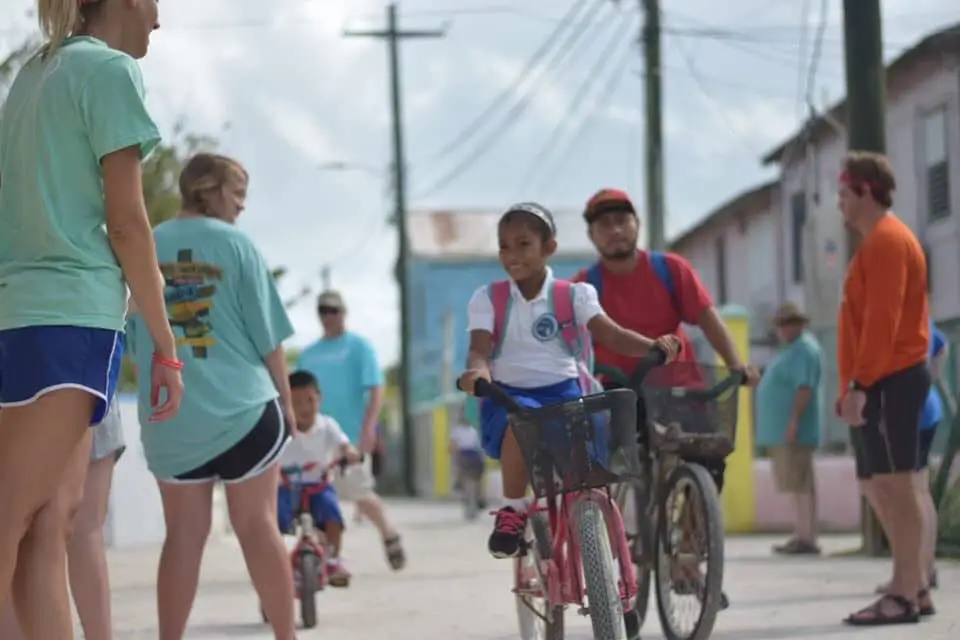 Children biking on a sunny street with onlookers.